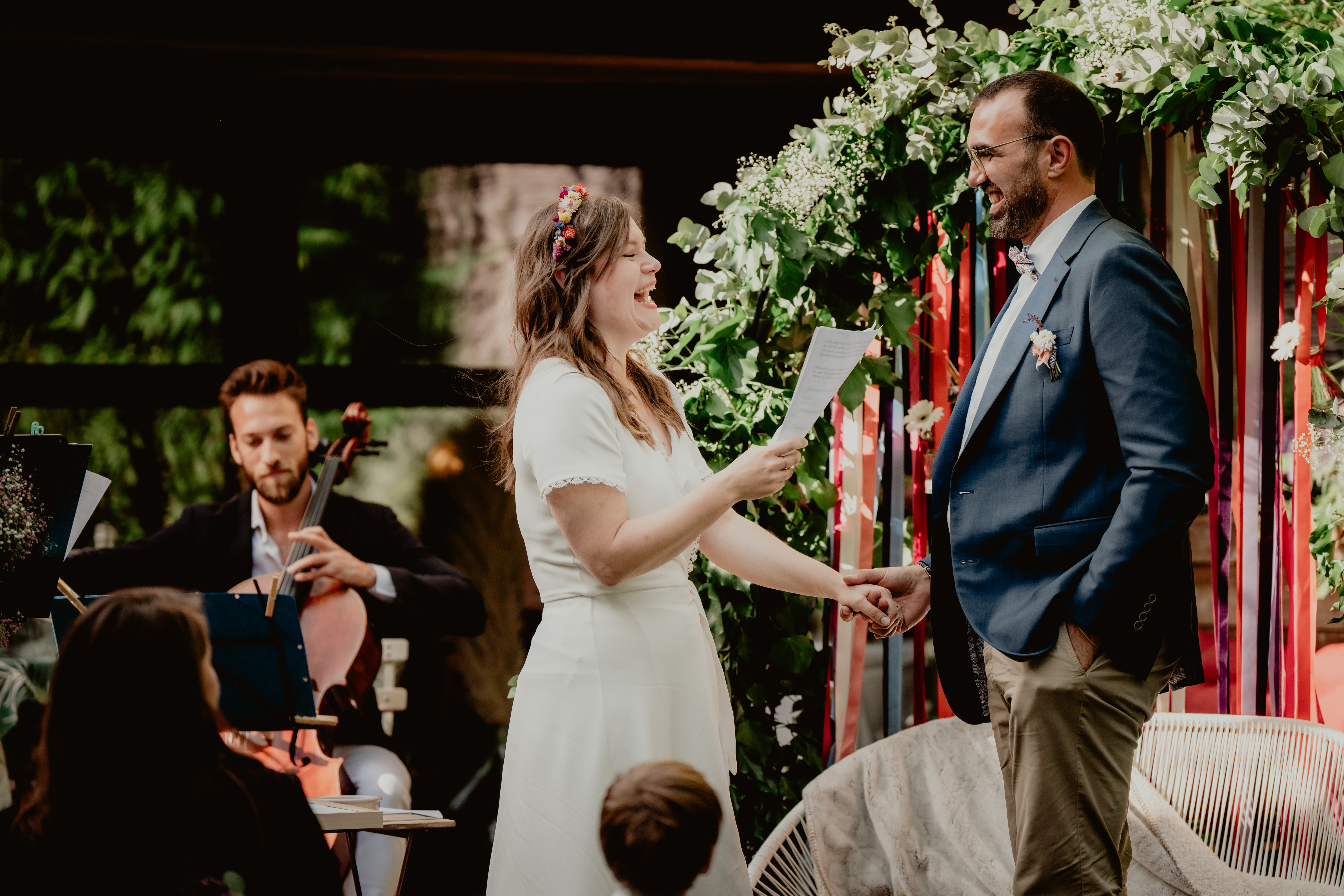 La mariée lit ses vœux face au marié pendant une cérémonie en extérieur, accompagnée d’un violoncelliste.