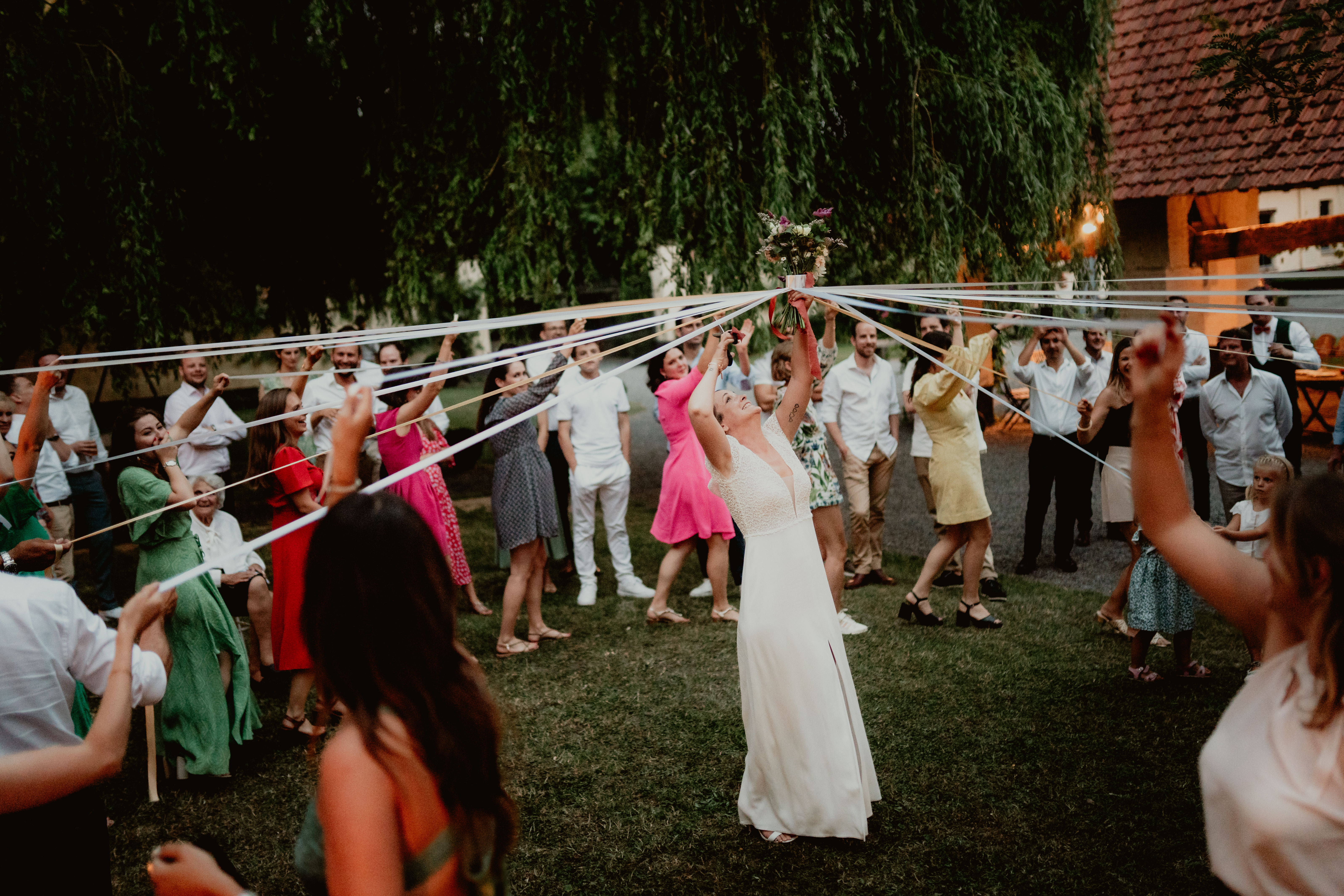 La mariée lève son bouquet au centre des invités qui tiennent des rubans.
